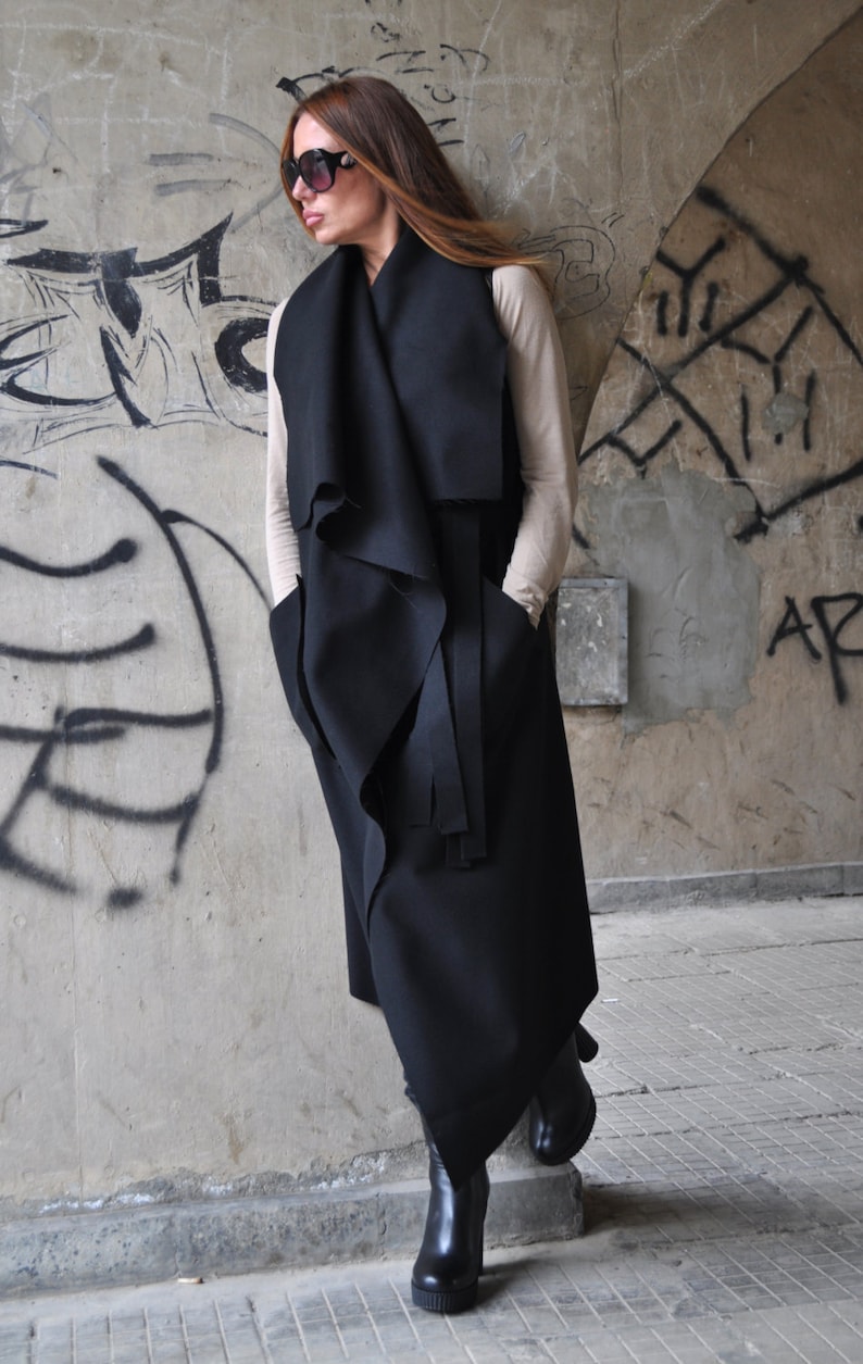 May include: A black long-sleeved wrap vest with a draped design and a tie belt. The vest is worn over a beige top and black boots.