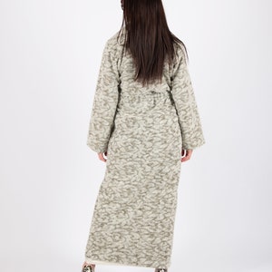 May include: A long, textured coat in shades of gray and beige, with a subtle pattern. The coat has a belted waist and wide sleeves. The person is wearing patterned high heels. The coat is a stylish outerwear option.
