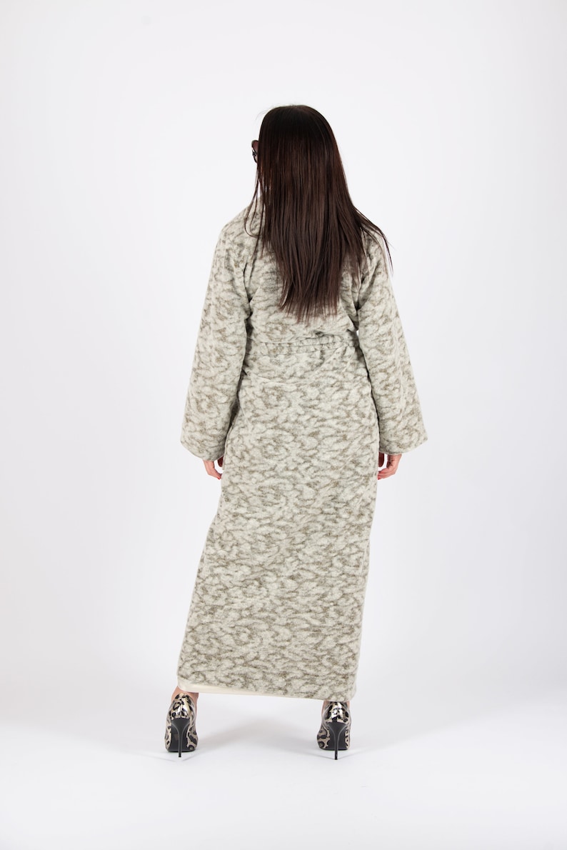 May include: A long, textured coat in shades of gray and beige, with a subtle pattern. The coat has a belted waist and wide sleeves. The person is wearing patterned high heels. The coat is a stylish outerwear option.