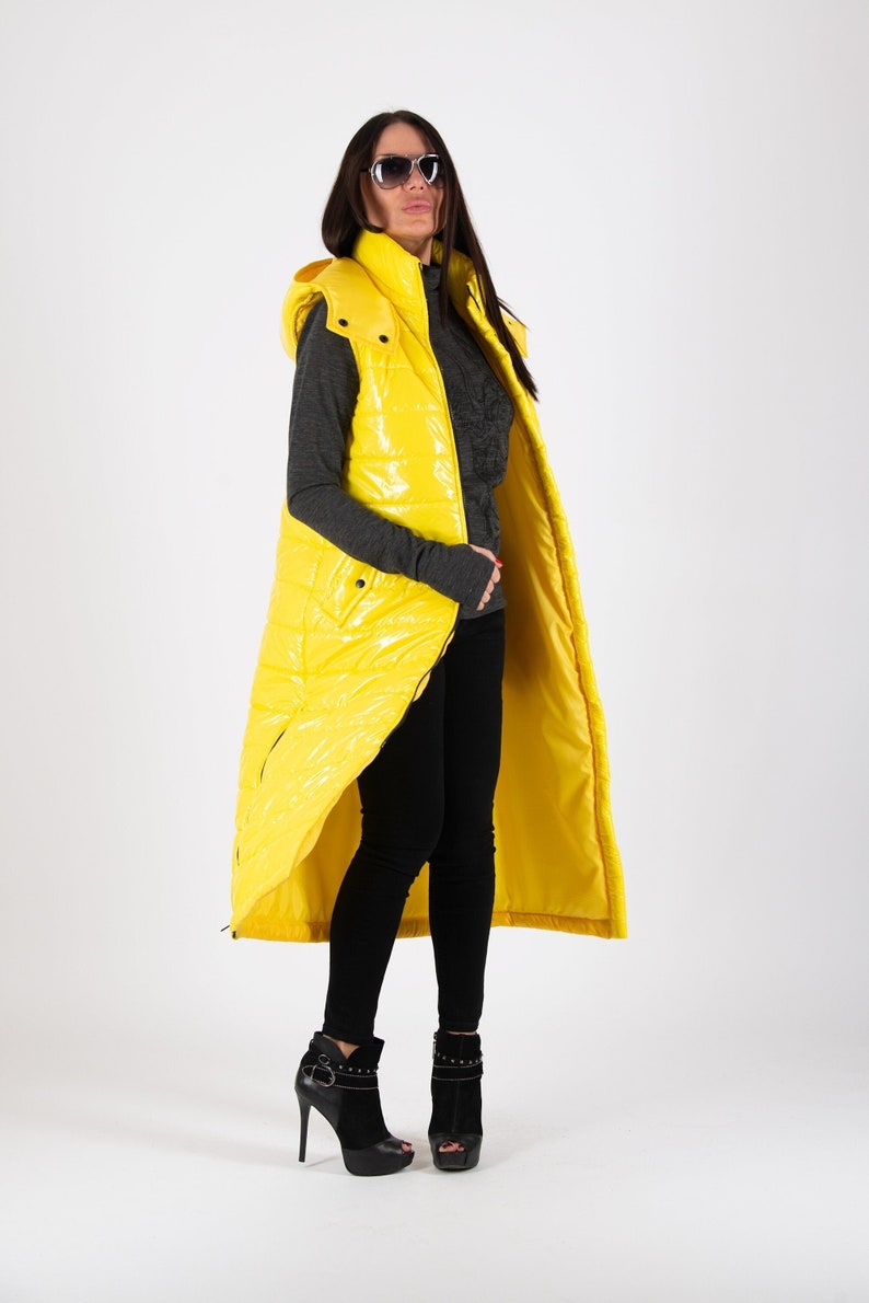 May include: A long yellow puffer vest with a hood. The vest is shiny and has a zipper closure. The model is wearing black pants and black heeled shoes.