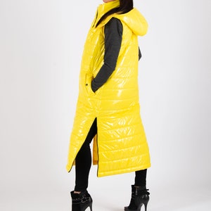 May include: A long yellow puffer vest with a hood. The vest has a zipper down the front and a zipper on each side. The model is wearing black pants and black heeled boots.