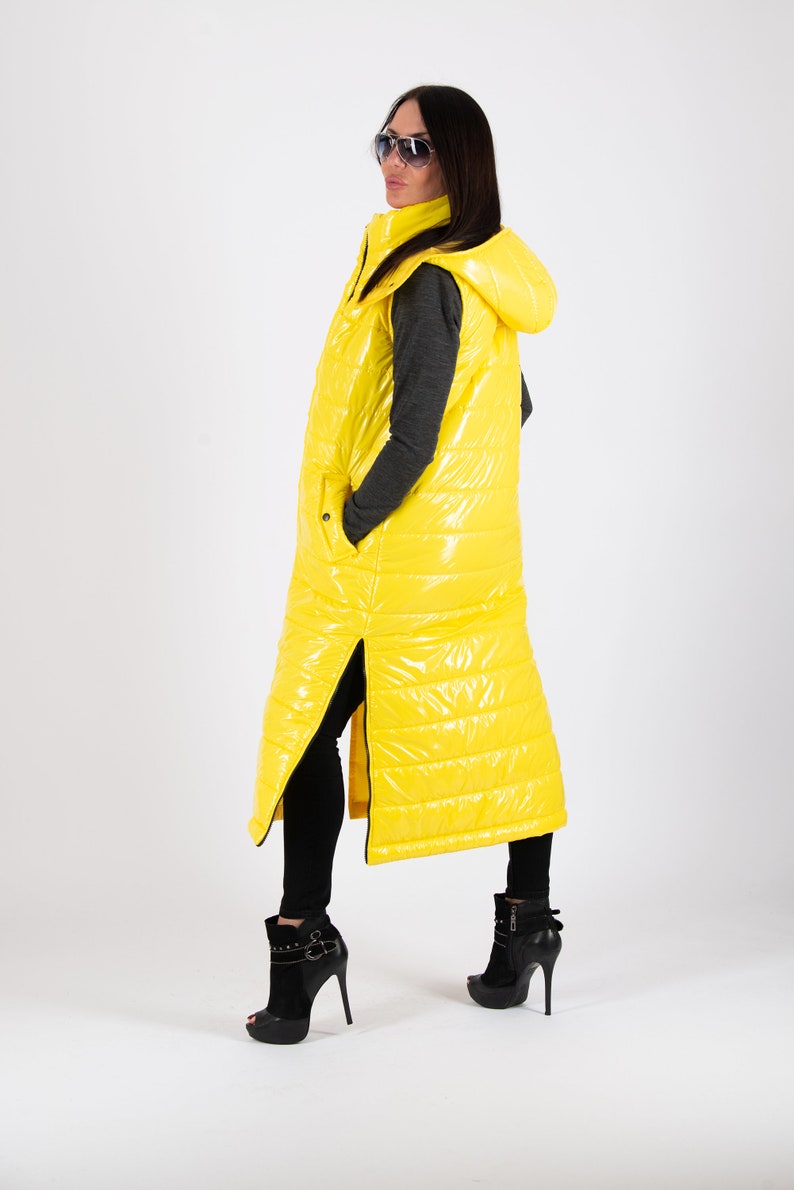 May include: A long yellow puffer vest with a hood. The vest has a zipper down the front and a zipper on each side. The model is wearing black pants and black heeled boots.
