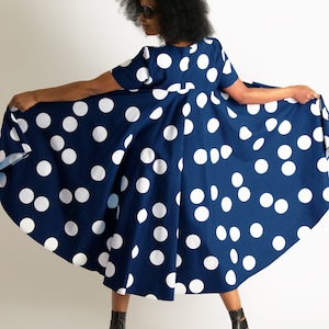 May include: A navy blue dress with large white polka dots. The dress has short sleeves and a flared skirt. The person in the image is wearing black high-heeled sandals. The dress is being held out to display its full shape.