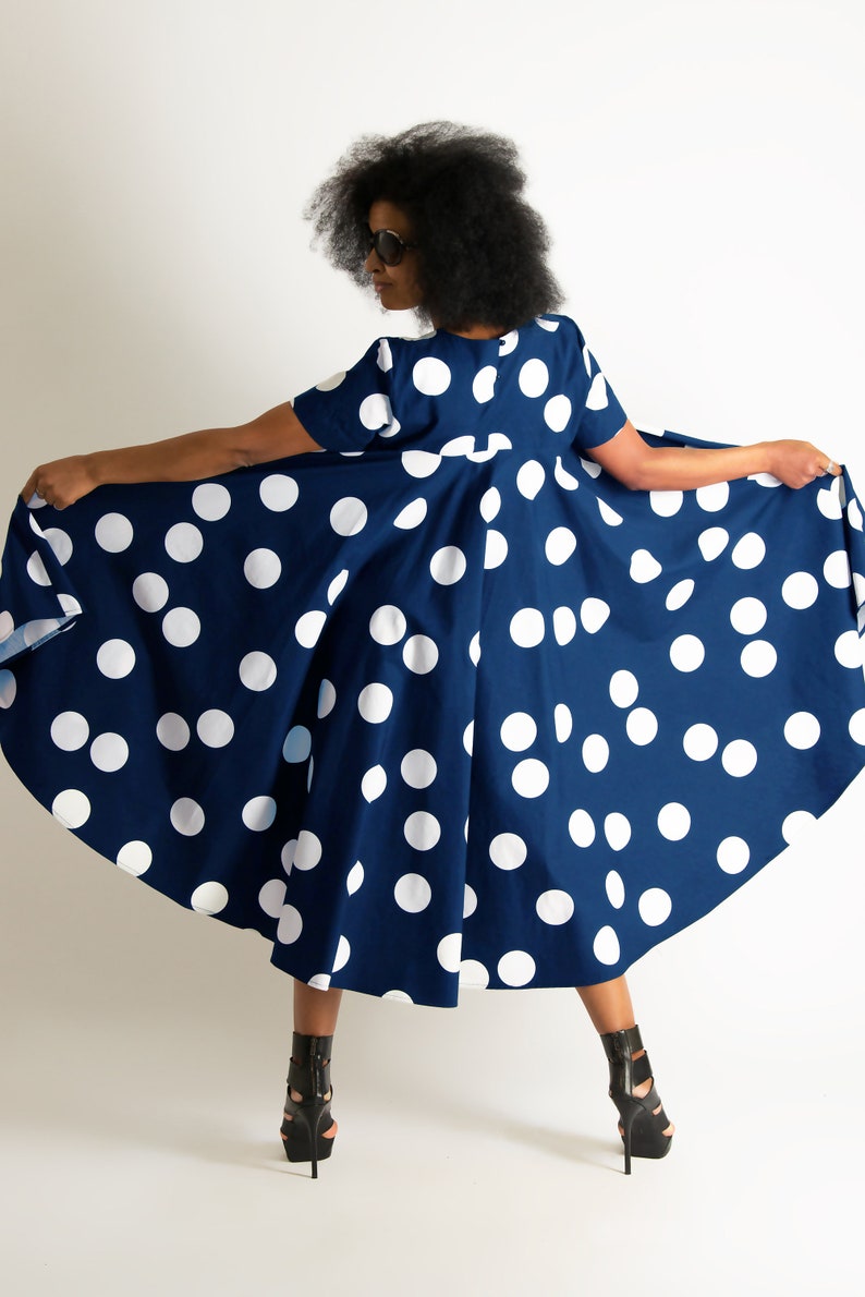 May include: A navy blue dress with large white polka dots. The dress has short sleeves and a flared skirt. The person in the image is wearing black high-heeled sandals. The dress is being held out to display its full shape.
