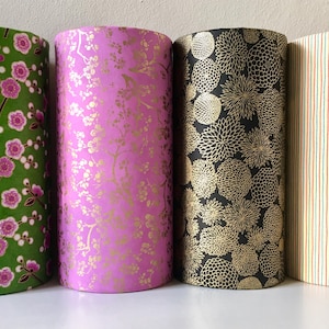 May include: Four cylindrical containers with decorative paper wraps. The containers feature floral and geometric patterns in green, pink, black, and cream. The designs include pink flowers, gold floral, and vertical stripes.