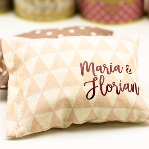 May include: A light pink pillow with a white triangle pattern and the text "Maria & Florian" in maroon lettering.