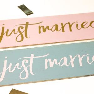 以下が含まれることがあります： 「just married」と書かれた木製の看板が2つ。上の看板はピンクで金色の文字、下の看板は薄い青で白い文字です。