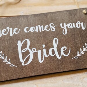 Könnte beinhalten: Ein rustikales Holzschild mit dem Text "here comes your bride" in weißer Kursivschrift. Das Schild ist mit weißen, blattförmigen Zweigen auf beiden Seiten des Textes verziert.