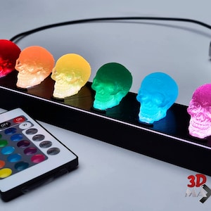 Puede incluir: Un conjunto de seis luces LED de colores con forma de calavera sobre una base negra con un mando a distancia. Las calaveras son rojas, naranjas, amarillas, verdes, azules y rosas.