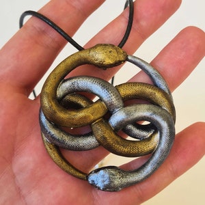 Könnte beinhalten: Eine Halskette mit einem Anhänger in Form eines goldenen und silbernen Schlangenknotens. Der Anhänger ist ein Knoten aus zwei Schlangen, eine golden und eine silber, die miteinander verschlungen sind. Die Halskette besteht aus einem schwarzen Band.