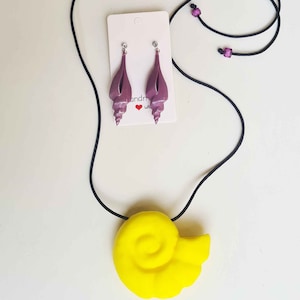 May include: A yellow seashell necklace with a purple seashell pendant. The necklace is on a black cord and the pendant is a 3D seashell.