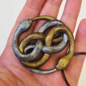 以下が含まれることがあります： 銀と金の蛇の結び目のペンダントネックレス。ペンダントは、結び目の形に絡み合った2匹の蛇でできています。蛇は銀と金で、ペンダントは黒いコードに取り付けられています。