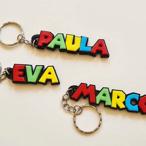 Puede incluir: Tres llaveros coloridos con los nombres "Paula", "Eva" y "Marcos" escritos en letras 3D. Cada letra es de un color diferente, incluyendo rojo, amarillo, verde y azul.