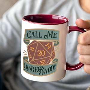 Puede incluir: Taza de cerámica blanca con interior y asa burdeos. La taza presenta el texto "CALL ME DUNGEON DADDY" con un gráfico de dado de veinte caras marrón y dorado. Un regalo divertido para los jugadores de juegos de mesa.