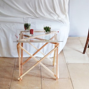 May include: A small, clear glass table with a wooden frame. The table has a simple, modern design and is perfect for a small space. The table is made of natural wood and has a clear glass top. The table is sitting on a light wood floor.