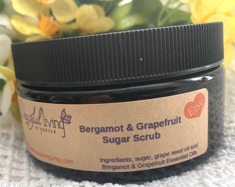 May include: A black jar with a brown label that reads "Bergamot & Grapefruit Sugar Scrub". The label also lists the ingredients: sugar, grape seed oil, and bergamot & grapefruit essential oils. The jar is sitting on a white crocheted surface.