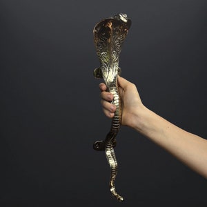 May include: A gold-toned door handle shaped like a cobra with intricate details. The handle is held in a hand against a dark gray background.