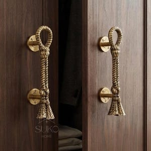 May include: Two brass door handles with a rope-like texture and tassel design mounted on dark wood cabinet doors. Each handle features a circular mounting plate with visible screws and a vertical rope motif that ends in a decorative tassel detail.