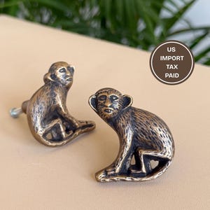 May include: Two antique brass-toned metal drawer knobs shaped like seated monkeys. Each knob features a textured finish and a screw attachment on the back. A circular brown sticker with the white text US IMPORT TAX PAID is visible in the background.