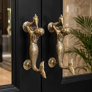 Peut inclure: Deux poignées de porte en laiton en forme de sirène, montées sur une porte noire. Les sirènes sont détaillées avec des écailles et des cheveux flottants, tenant leurs bras au-dessus de leur tête. Les poignées ajoutent une touche décorative à l'entrée.