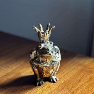 May include: A gold frog figurine wearing a crown, sitting on a wooden surface.