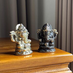 May include: Two small, ornate, brass elephant statues. One is gold and the other is dark brown. They are both sitting on a wooden surface.