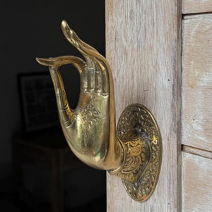 Könnte beinhalten: Ein goldfarbener Türgriff aus Messing in Form einer Hand in einer Willkommensgeste. Der Griff ist aufwendig mit floralen Mustern verziert und an einer Holztür montiert.