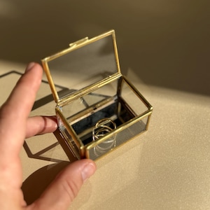 May include: A small, rectangular glass jewelry box with a gold-colored metal frame. The box is open, revealing two gold rings inside. The box is held by a hand, and the background is a neutral beige color.