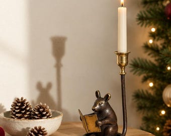 May include: A bronze-toned metal candle holder featuring a sculpted mouse reading a book, with a tall, slender stem supporting a brass-colored cup holding a single white taper candle. The base is circular and supports the mouse figure.
