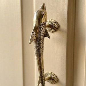 May include: A brass door handle shaped like a fish, with detailed scales and fins. The handle is mounted on a cream-colored door, with decorative floral backplates. The fish design adds a unique, nautical touch to the door.
