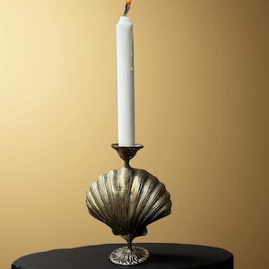 May include: A white candle with a flame burning on top is placed in a gold metal seashell-shaped candle holder. The candle holder is on a black surface.