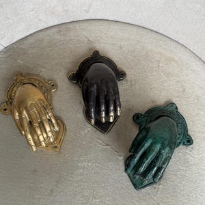 May include: Three antique hand-shaped door knockers in gold, black, and green. Each knocker features a detailed hand design with a decorative backplate. The gold knocker has a bright finish, while the black and green knockers have a darker, aged appearance.