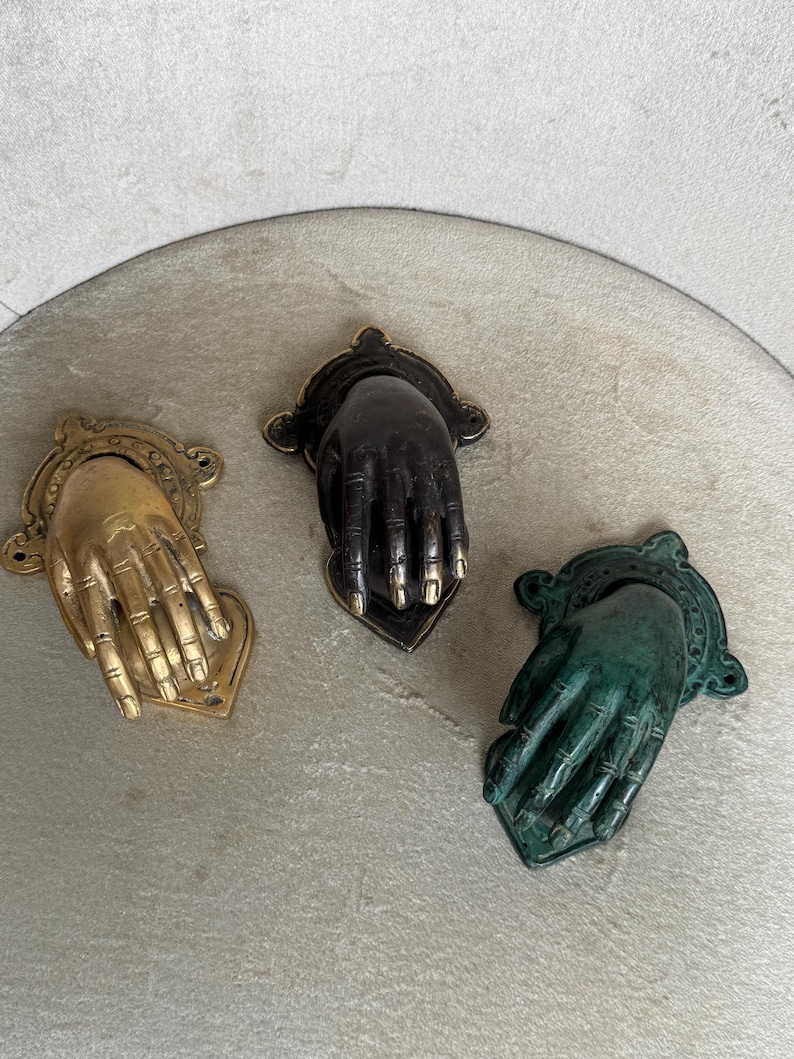 May include: Three antique hand-shaped door knockers in gold, black, and green. Each knocker features a detailed hand design with a decorative backplate. The gold knocker has a bright finish, while the black and green knockers have a darker, aged appearance.