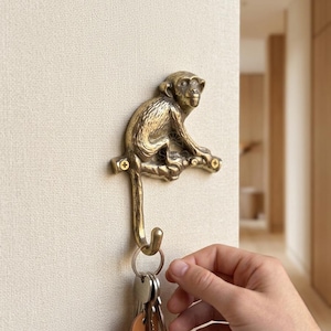 Könnte beinhalten: Ein messingfarbener Wandhaken aus Metall in Form eines auf einem Ast sitzenden Affen. Der Affe ist in sitzender Position dargestellt, wobei sein Schwanz nach unten ragt und einen Haken bildet. Zwei goldfarbene Schrauben befestigen den Ast.