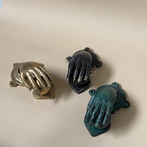 May include: Three decorative hand-shaped door knockers in brass, black, and green. Each knocker features a detailed hand design with a heart-shaped base. The brass knocker has a vintage look, while the black and green knockers have a more modern finish.
