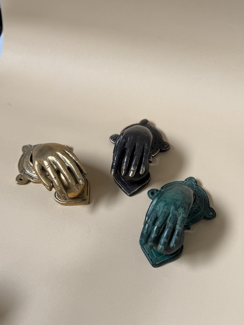 May include: Three decorative hand-shaped door knockers in brass, black, and green. Each knocker features a detailed hand design with a heart-shaped base. The brass knocker has a vintage look, while the black and green knockers have a more modern finish.