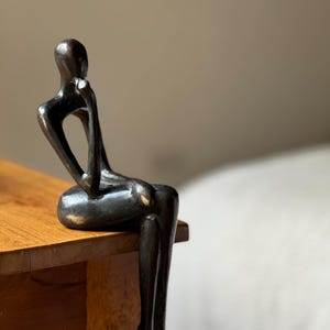 May include: A black metal sculpture of a person sitting with their legs dangling off the edge of a wooden surface.