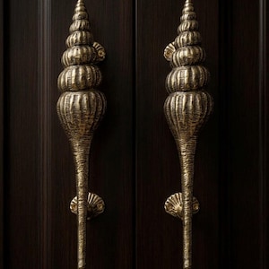 May include: Two ornate, brass-colored door handles. Each handle is shaped like a stylized seashell, with a spiraling top and a long, textured stem. They are mounted on a dark brown wooden door.