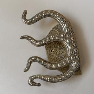 May include: A silver-toned octopus wall hook. The hook has three curved tentacles emerging from a textured, circular base. The design has a detailed, antique appearance, suitable for nautical or coastal decor.