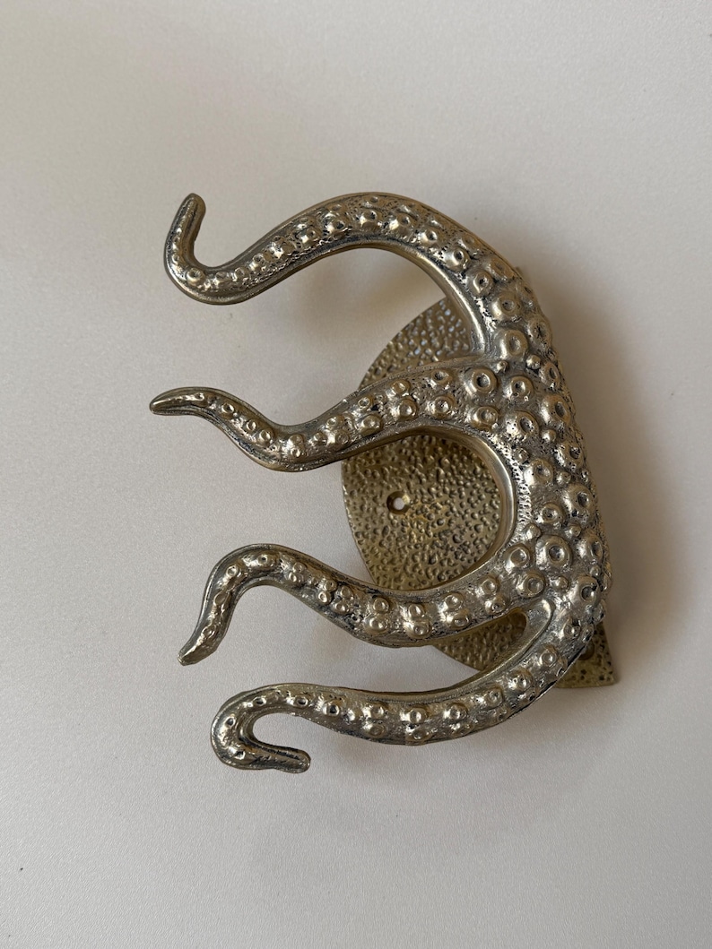 May include: A silver-toned octopus wall hook. The hook has three curved tentacles emerging from a textured, circular base. The design has a detailed, antique appearance, suitable for nautical or coastal decor.