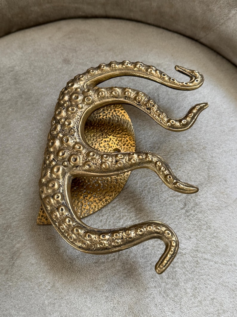 May include: A brass-coloured, octopus-shaped decorative object. The object has a textured surface with multiple tentacles extending outwards. The base is a textured, heart-shaped design. It is likely a decorative door handle or wall hanging.
