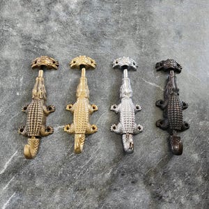 Pode incluir: Quatro ganchos de parede de metal em forma de jacaré dispostos em fila. Cada gancho apresenta um corpo reptiliano texturizado com uma cauda curva na parte inferior e uma pequena placa de montagem arredondada no topo. Latão, ouro, prata e bronze.