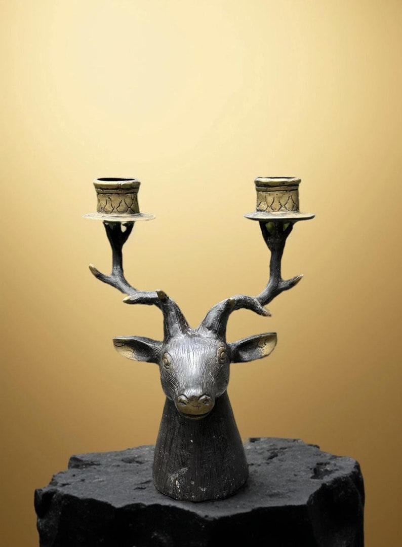 May include: A black deer head candle holder with two gold candle cups. The deer head is detailed with a textured finish and the candle cups have a decorative pattern.