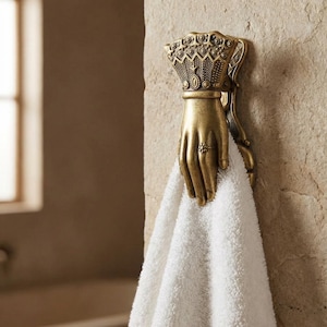 Puede incluir: Un colgador de toallas dorado en forma de mano con detalles intrincados, montado en una pared de piedra texturizada. Una toalla blanca y esponjosa cuelga de él. La mano presenta un anillo y un diseño similar a una corona.
