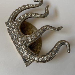 May include: A silver-coloured metal wall hook in the shape of an octopus tentacle. The hook features a textured surface with small, round details and is mounted on a decorative backplate. The tentacles curve upwards, creating a unique and functional design.