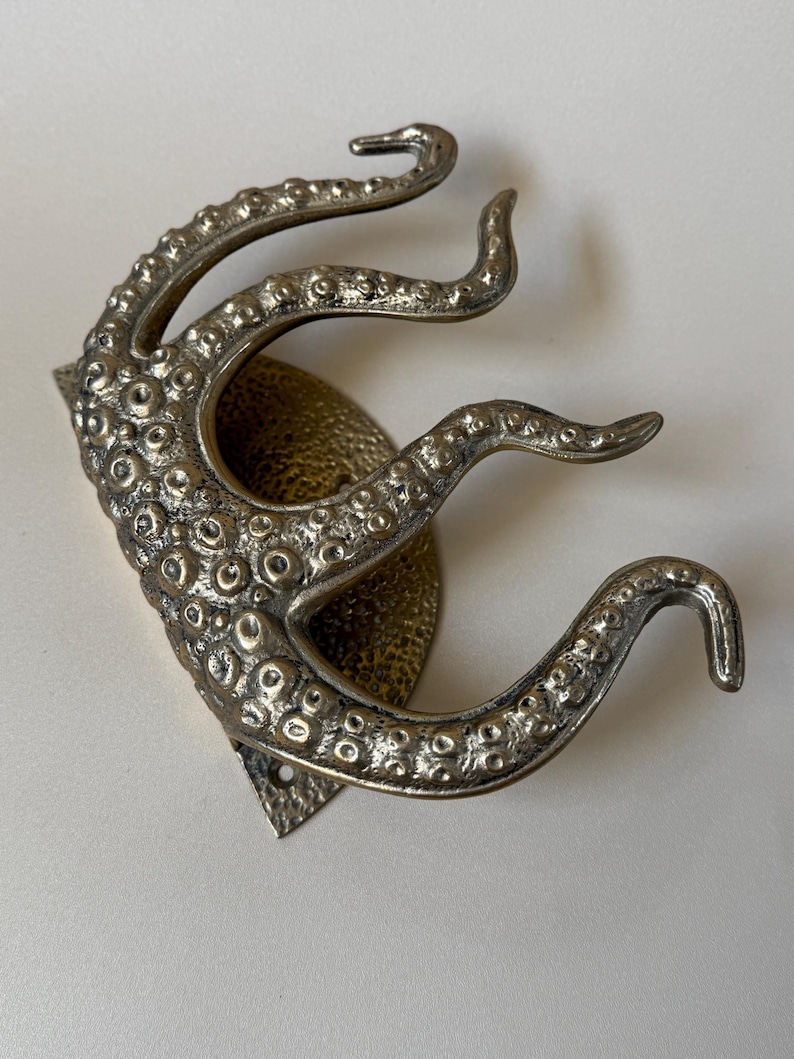 May include: A silver-coloured metal wall hook in the shape of an octopus tentacle. The hook features a textured surface with small, round details and is mounted on a decorative backplate. The tentacles curve upwards, creating a unique and functional design.