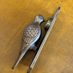May include: A brass door knocker shaped like a bird with detailed wing feathers and a smooth head. The bird is attached to a long, rectangular metal mounting plate that features a circular strike plate at the top for installation on a door.