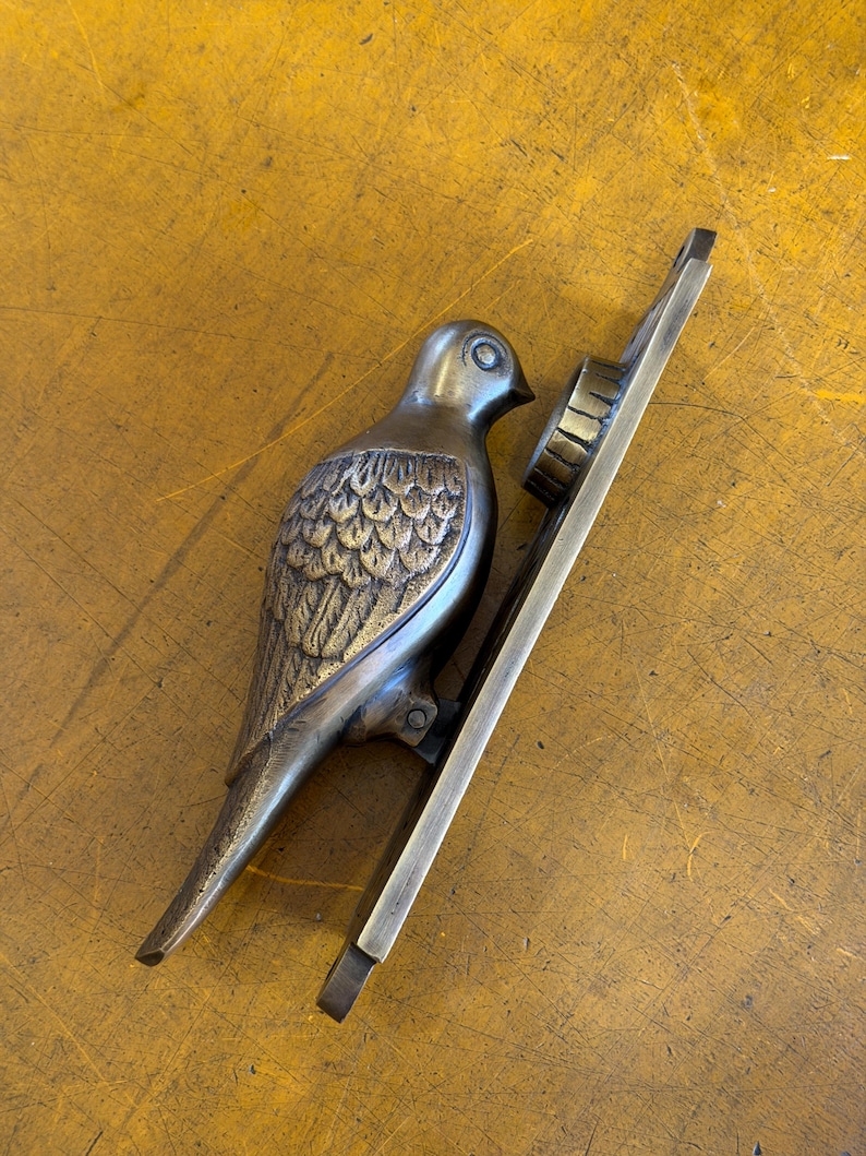 May include: A brass door knocker shaped like a bird with detailed wing feathers and a smooth head. The bird is attached to a long, rectangular metal mounting plate that features a circular strike plate at the top for installation on a door.