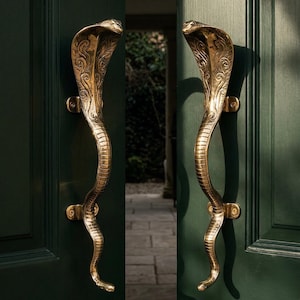 May include: A pair of ornate brass door handles shaped like cobras. The handles are mounted on a dark green door, with intricate detailing on the cobra's head and body. The door is slightly ajar, revealing a glimpse of an outdoor pathway.