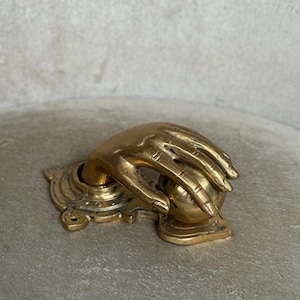 May include: A brass door knocker in the shape of a hand grasping a sphere. The hand is detailed with visible knuckles and lines, mounted on an ornate base. The finish is a warm, aged gold.